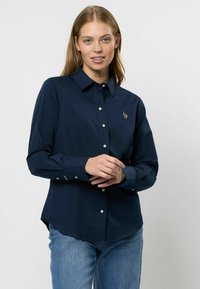 Tengerészkék gombos ing, gallérral, hosszú ujjal és fehér gombokkal. Kicsi hímzett mintával rendelkezik a mellrészén. Lazább szabású.