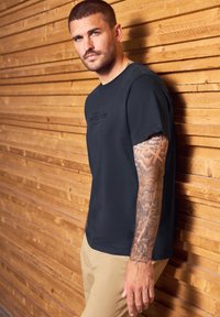 Muž s krátkými vlasy a tetovanou pravou paží, oblečený v černém tričku a béžových kalhotách, opírá se o dřevěnou stěnu a dívá se vpřed.