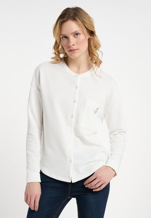 Camisa blanca de manga larga con botones y cuello redondo, con un bolsillo en el pecho y una textura de tela suave y lisa.