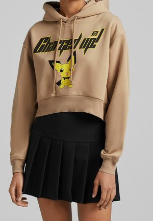 Personne portant un sweat à capuche court beige avec "Charged up!" et un motif Pikachu, associé à une jupe plissée noire sur un fond uni.