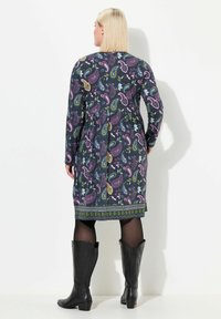 Långärmad klänning i mörkt mönstrad tyg med paisley- och blommönster i lila, grönt och gult, kombinerad med svarta cowboyboots.
