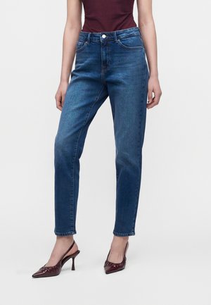 Donna che indossa jeans di denim blu a vita media e scarpe slingback con tacco color bordeaux, in piedi con le mani rilassate lungo i fianchi.