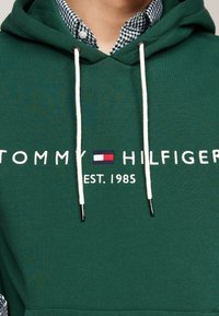 Zelená mikina s bílými šňůrkami, na které je logo s nápisem "TOMMY HILFIGER" a "EST. 1985" v bílém textu. Nahoře oblečená přes kostkovanou košili.