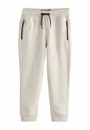 Beżowe spodnie dresowe z elastycznym pasem, z troczkami do regulacji, oraz dwoma bocznymi kieszeniami na zamek; ściągacze przy kostkach.