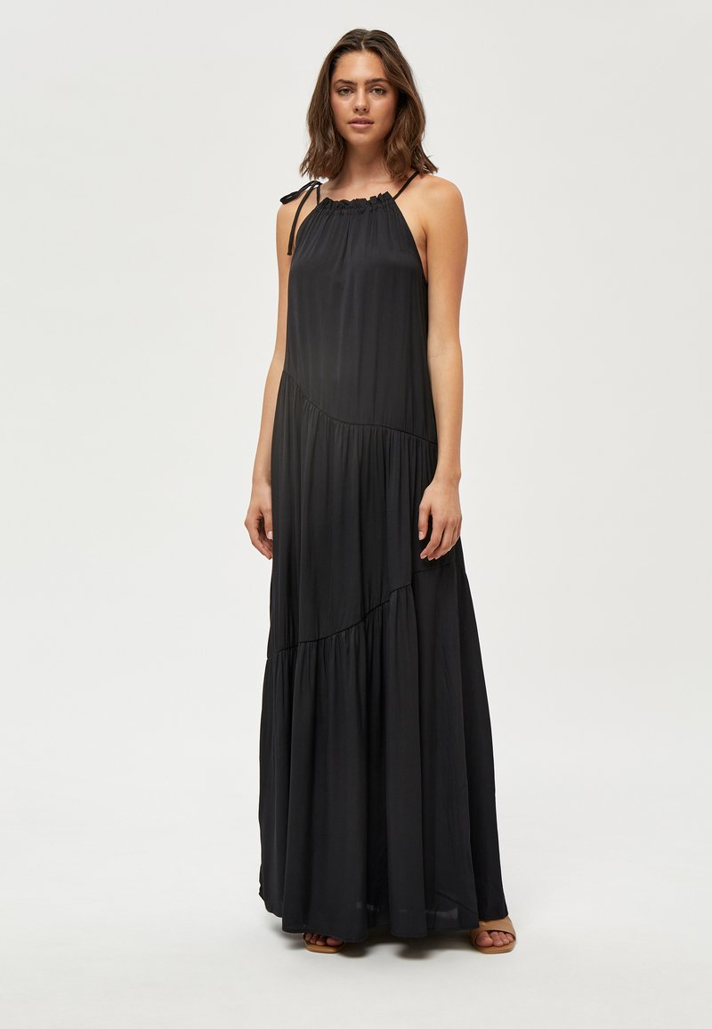 Naine seisab, kandes varrukateta musta maksikleit, millel on asümmeetrilised kihid ja õhukesed õlapaelad, taustal on lihtne hele taust.