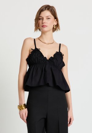 Giovane donna che indossa un top nero senza maniche con balze, pantaloni neri, collana d'oro, orecchini ad anello e diversi braccialetti d'oro al polso sinistro.