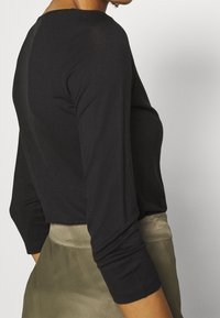 Una persona è vista di lato, indossando un top nero aderente a maniche lunghe e una gonna chiara, con le mani appoggiate nelle tasche.