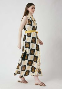 Long, sleeveless dress featuring a floral pattern in yellow, black, and white, with a yellow belt at the waist and side slits.