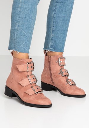 Roze suède enkellaarsjes met drie brede banden en metalen gespen, lage blokhak en een zijrits. Gedragen met versleten denim.