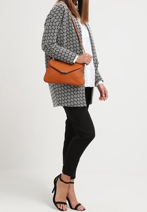 Kobieta w czarno-białej marynarce w wzory, białej koszuli, czarnych spodniach, czarnych szpilkach, trzymająca brązową torebkę listonoszkę.