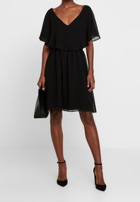 Black dress with a V-neck, sheer flutter sleeves, an elastic waist, and a flowy skirt. Paired with a black handbag and heeled shoes.