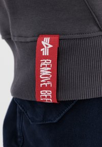 Felpa grigia con polsini a coste, che presenta un'etichetta rossa prominente con la scritta "RIMUOVI PRIMA" in lettere bianche. Tessuto blu navy visibile sotto.