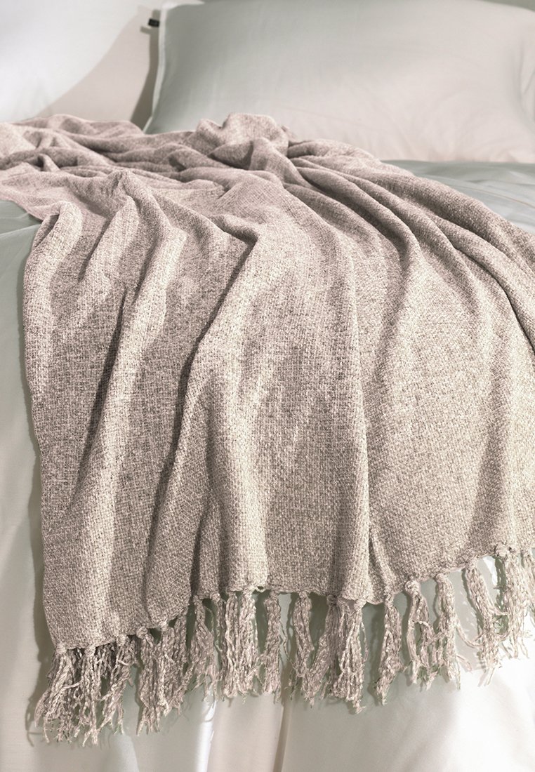 Texturovaná šedá deka s třásněmi, přehozená přes světlé povlečení, ukazující měkkou tkaninu a uvolněný design.