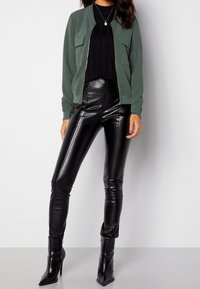 Giacca bomber verde con due tasche frontali, pantaloni in ecopelle neri slim-fit e stivaletti neri con punta. Texture lisce, design aderente.