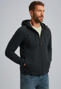Zwarte hoodie met ritszakken, textuurstof, voorkeursritssluiting en verstelbare trekkoorden. Draagt over een lichtgekleurde shirt.
