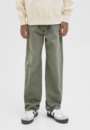 Personne portant un pantalon ample vert olive, un sweat crème et des baskets blanches avec des rayures noires, debout sur un fond uni.