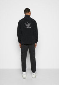 Svart fleecejacka med hög krage och vitt Adidas-logotyp på ryggen, kombinerad med svarta mjukisbyxor och vita sneakers.