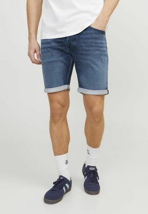 Shorts en denim dans un bleu foncé avec des ourlets retroussés, associés à des chaussettes blanches et des baskets sombres. Le design présente une coupe classique et des poches.