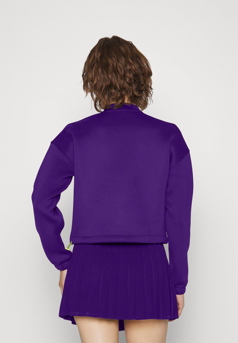 Sweatshirt violet à manches longues et poignets côtelés, associé à une jupe violette plissée. Texture lisse et couleur vive.