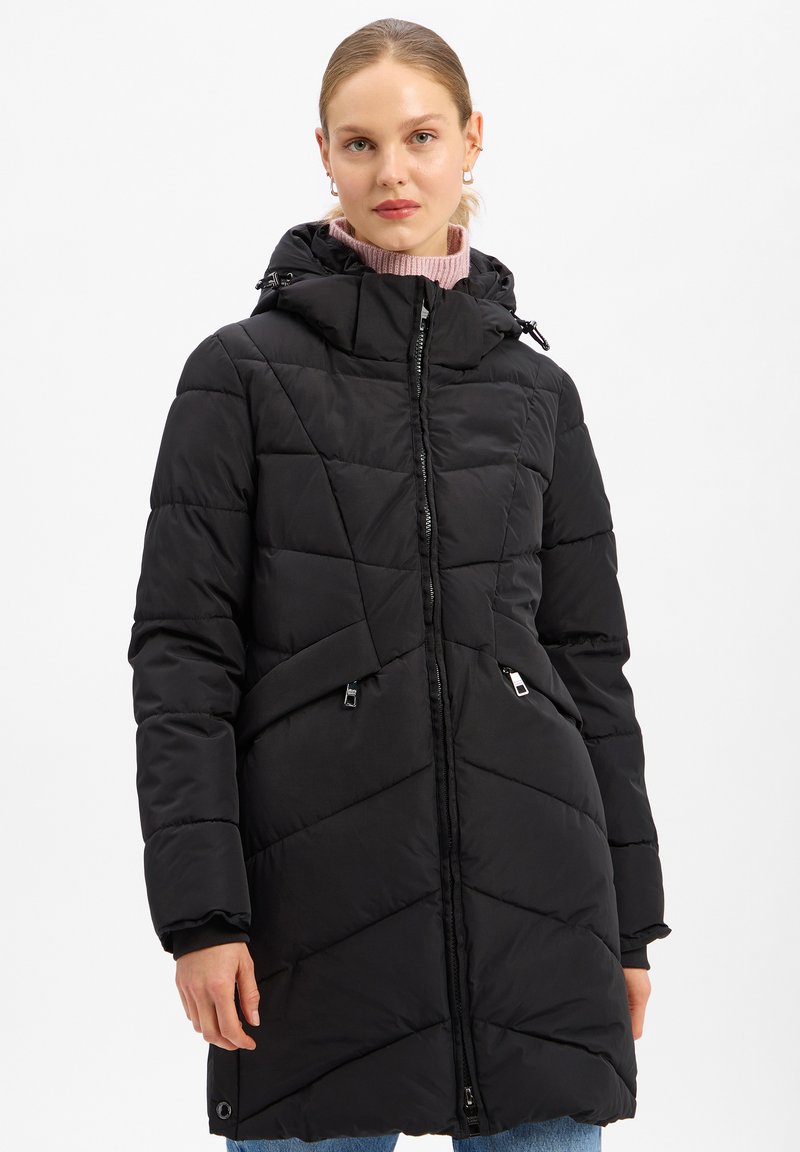 Vrouw in een zwarte gewatteerde winterjas met capuchon over een roze coltrui en blauwe spijkerbroek, staande tegen een witte achtergrond.