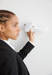 Vit sneaker med ett perforerat mönster, med en guldlogotyp och grå accenter, hålls uppe av en person i en svart kavaj.