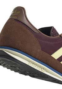 Bruna mockaskinnsneakers med vinröd tyg, gula ränder, bakdel i brunt läder och svart gummisula med texturerat mönster.