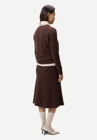 Femme portant un pull en maille marron et une jupe midi assortie, une chemise blanche à col en dessous, des chaussettes blanches froissées et des chaussures blanches à talons.
