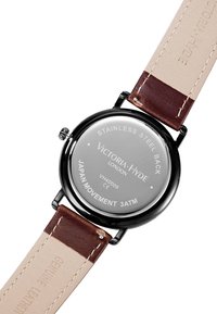 Montre ronde avec un fond en acier inoxydable, un bracelet en cuir véritable marron, et un cadran argenté. Le texte gravé comprend des détails sur la marque et le mouvement.
