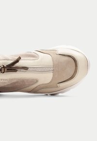 Zapatilla de cuero y ante beis con un acabado texturizado, detalle de cremallera y suela de goma blanca. Presenta un diseño aerodinámico con secciones contrastantes.