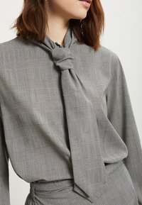 Siva karirana bluzica sa čvorom na ovratniku, izrađena od lagane tkanine s glatkom teksturom i suptilnim sjajem.