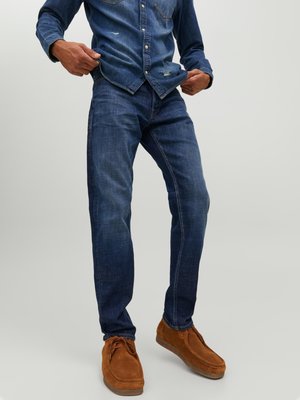 Jeans de mezclilla azul oscuro con un diseño ajustado, ligeras desgastes y una textura suave, acompañados de zapatos de ante marrón.