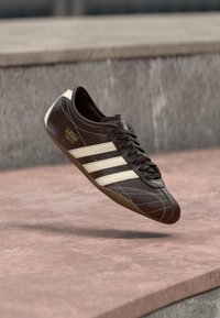 Sapatilha de couro castanho com riscas creme, biqueira redonda, superfície texturizada e sola de borracha cor de goma, com a marca "adidas Tokyo" na lateral.