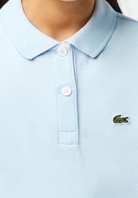 Světlomodré polotričko z texturované látky, s dvěma knoflíky u límce a malým zeleným vyšívaným krokodýlím logem na hrudi.