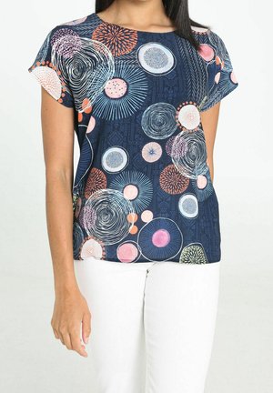 Kobieta w granatowej bluzce z krótkim rękawem z kolorowymi okrągłymi wzorami, w połączeniu z białymi spodniami, stojąca na jednolitym tle.