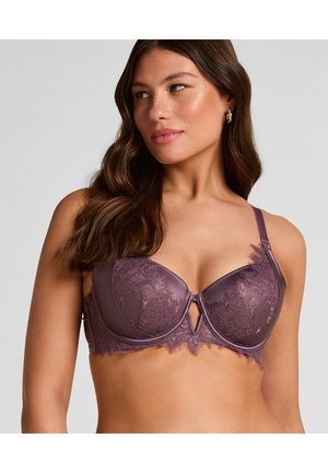 Woman with long brown hair wearing a purple lace and satin bra, looking to the side against a plain light background.