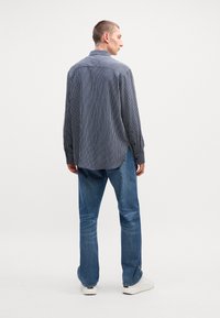 Ριγέ μακρυμάνικο πουκάμισο σε ναυτικό μπλε και λευκό, με ελαφρώς καμπυλωτό στρίφωμα και κουμπωτά μανίκια, συνδυασμένο με ανοιχτό μπλε τζιν και λευκά αθλητικά παπούτσια.