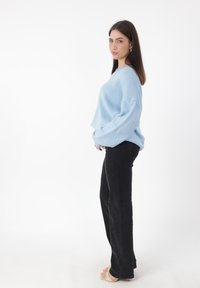 Hellblaues oversized Sweatshirt mit strukturiertem Finish, kombiniert mit schwarzen, ausgestellten Jeans und beigen High Heels. Das Model posiert seitlich vor einem weißen Hintergrund.