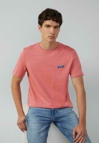 Młody mężczyzna z kręconymi ciemnymi włosami, ubrany w różowy t-shirt w kolorze łososiowym z małym niebieskim emblematem samochodu oraz jasnoniebieskie dżinsy, siedzi na tle jednolitego tła.