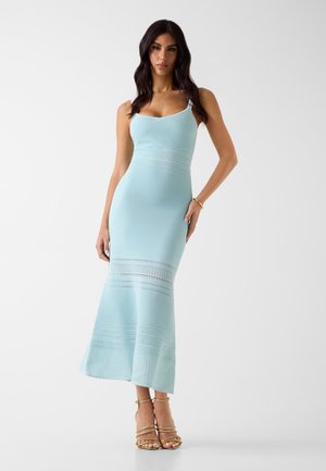 Frau trägt ein figurbetontes hellblaues Strickkleid mit dünnen Trägern und gemusterten horizontalen Streifen, kombiniert mit goldfarbenen, geschnürten High-Heel-Sandalen.