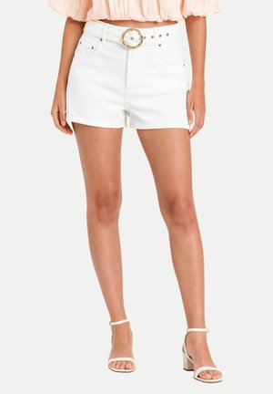 Donna che indossa pantaloncini bianchi a vita alta con cintura ad anello dorato, crop top pesca con maniche a sbuffo e sandali bianchi con tacco largo e cinturino alla caviglia.