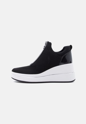 Baskets compensées en maille noire avec fermeture éclair frontale, semelle plateforme blanche haute, et panneau de talon noir brillant, vue latérale sur fond blanc.