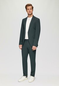 Dunkelgrüner Anzug mit schlanker Passform, getragen über einem weißen T-Shirt. Ein Paar weiße Sneaker vervollständigt das Outfit. Glatter Stoff, minimalistischer Stil.