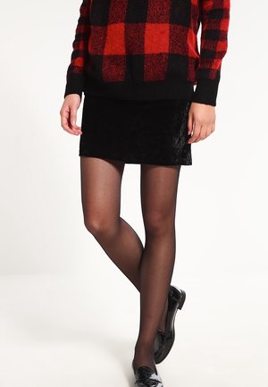 Personne portant un pull à carreaux rouge et noir, une mini-jupe noire, des collants noirs transparents et des mocassins noirs brillants, debout avec les jambes croisées.