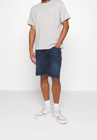 Šedé bavlněné tričko s krátkými rukávy a kulatým výstřihem, v kombinaci s modrými džínovými šortkami střední délky a bílými sportovními botami se šedými akcenty.