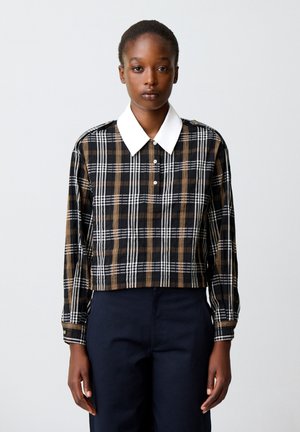 Donna con pelle scura che indossa una camicia a quadri nera, bianca e marrone con colletto bianco e pantaloni scuri, in piedi davanti a uno sfondo chiaro e uniforme.