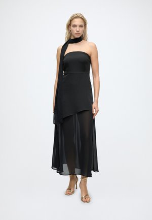 Mujer con un vestido negro sin tirantes con una superposición asimétrica y transparente y una bufanda a juego en el cuello, combinados con sandalias doradas de tacón alto.