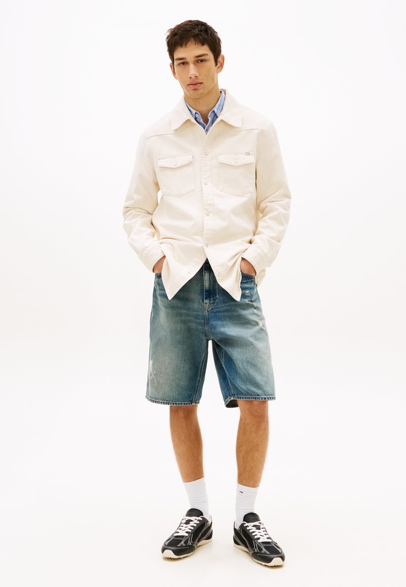 Jonge man met een lichtbeige overhemd met knopen over een blauwe shirt, spijkershorts, witte sokken en zwarte sneakers, staand met handen in de zakken.