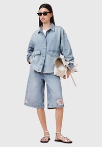 Giacca di jeans blu sbiadita con dettagli nelle tasche, abbinata a pantaloni Bermuda in denim strappato. Il modello tiene una borsa color crema e indossa occhiali da sole neri.