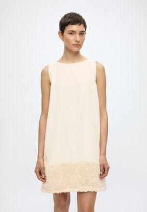 Γυναίκα με κοντά σκούρα μαλλιά που φοράει μπεζ αμάνικο φόρεμα με υφασμάτινη τελειώματα, στέκεται μπροστά σε ένα απλό φωτεινό φόντο.