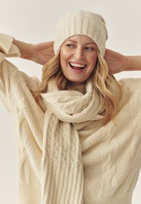 Set composto da berretto e sciarpa color crema in maglia con motivi a coste; indossato con un maglione spesso a trecce. Primo piano sulle trame e dettagli del design.